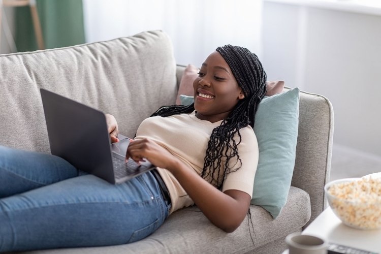 Joyful African American woman lounging on a couch smiling while looking at her laptop.